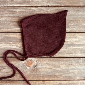 May include: A burgundy knit baby bonnet with long ties. The bonnet is made of a soft, textured material and is designed to cover the head and neck. The ties are used to secure the bonnet under the chin. The bonnet is displayed on a wooden surface.