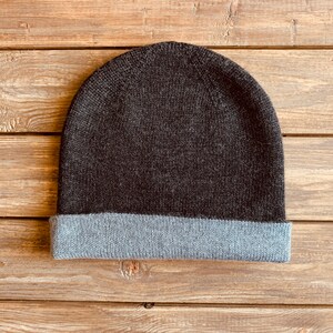 May include: A dark grey beanie with a light grey fold-over brim. The beanie is made of a soft, knitted fabric.