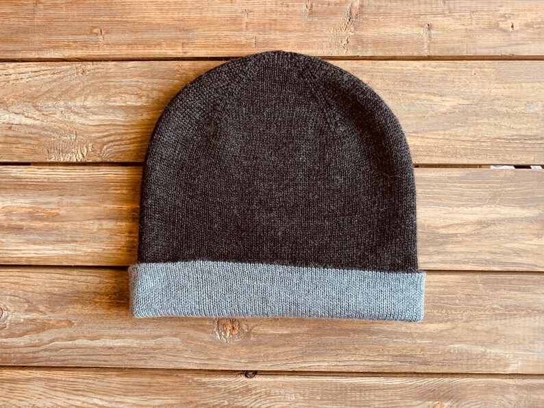 May include: A dark grey beanie with a light grey fold-over brim. The beanie is made of a soft, knitted fabric.