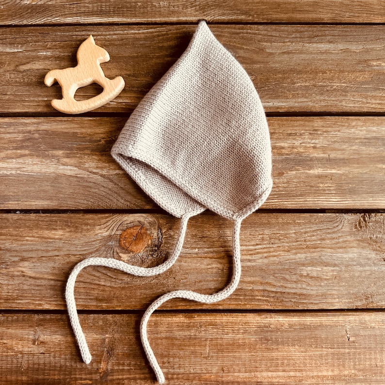 May include: A light gray knitted baby bonnet with long ties, lying on a wooden surface. A small wooden rocking horse toy is next to the bonnet. The bonnet is made of a soft, textured yarn.