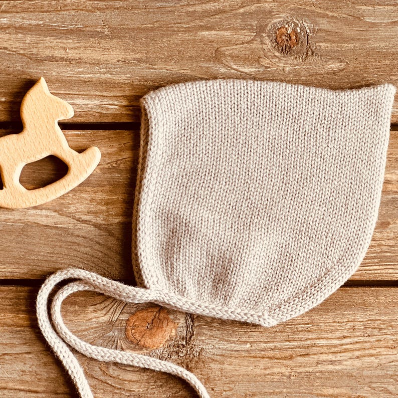 May include: A light beige knitted baby bonnet with ties, lying on a wooden surface. A wooden rocking horse toy is visible in the upper left corner. The bonnet is made of a soft, textured yarn.