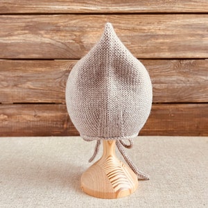 May include: A light gray knitted baby bonnet with a pointed top, displayed on a wooden stand. The bonnet has a textured appearance and ties under the chin with matching yarn. The background features a wooden plank wall.