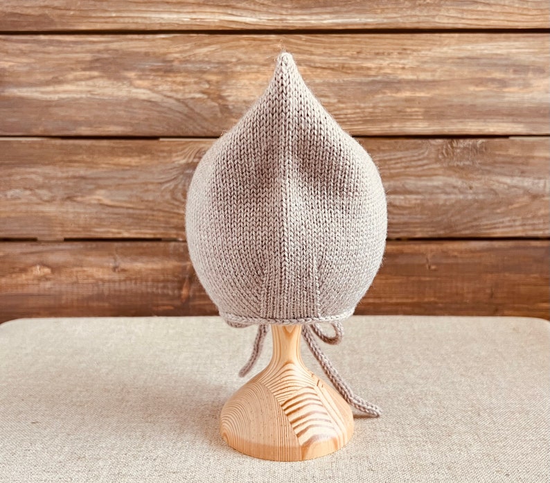 May include: A light gray knitted baby bonnet with a pointed top, displayed on a wooden stand. The bonnet has a textured appearance and ties under the chin with matching yarn. The background features a wooden plank wall.