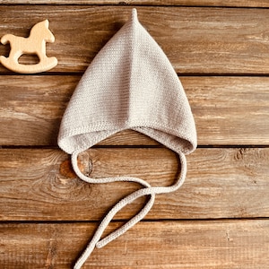 May include: A light gray knitted baby bonnet with a pointed top and long ties. A wooden rocking horse toy is to the left of the bonnet. The items are on a rustic wooden surface.