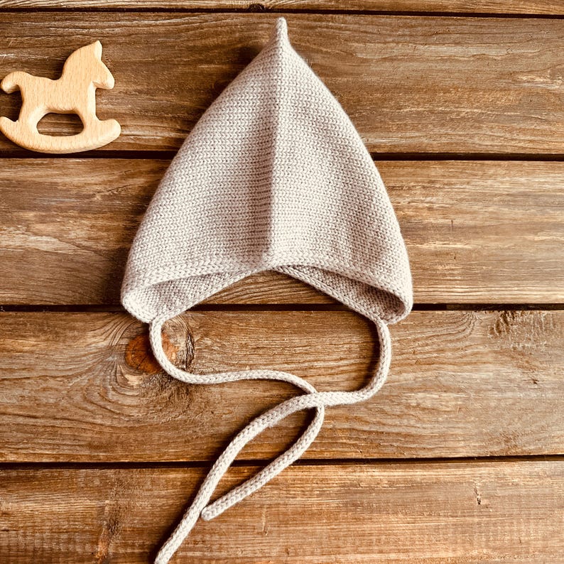 May include: A light gray knitted baby bonnet with a pointed top and long ties. A wooden rocking horse toy is to the left of the bonnet. The items are on a rustic wooden surface.