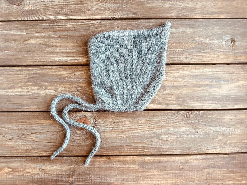 以下が含まれることがあります： 長い紐が付いた灰色のニットのベビーボンネット。ボンネットは柔らかくふわふわの糸でできており、先が尖っています。