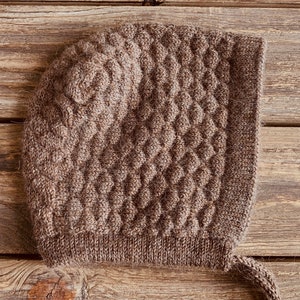 May include: A brown knitted baby bonnet with a textured, diamond-like pattern. The bonnet has a rounded top and a square brim, with a knitted tie on each side. The item is made of a soft, warm material.