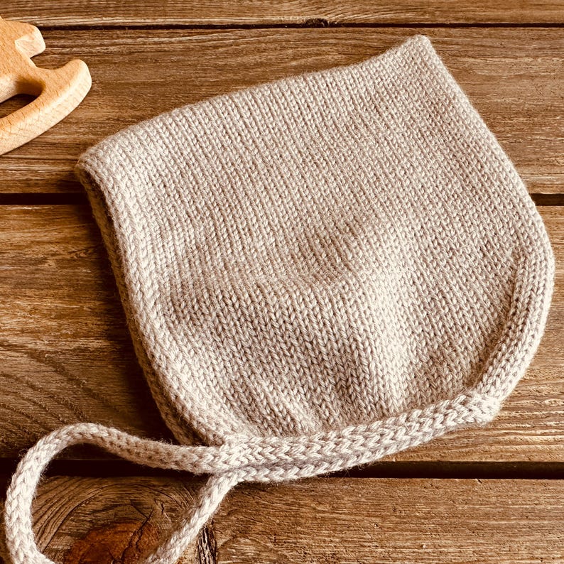 May include: A light beige knitted baby bonnet with tie strings. The bonnet is made of a soft, textured yarn and is set against a wooden background. The bonnet is designed to cover the head and tie under the chin.