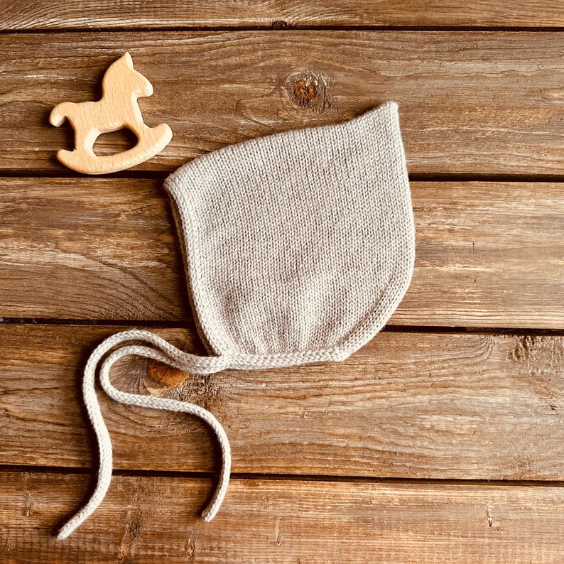 May include: A light gray knitted baby bonnet with tie strings, next to a small wooden rocking horse toy, set on a rustic wooden surface. The bonnet is made of a soft, textured yarn.