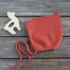 May include: A rust-colored knitted bonnet with a pointed top and two long matching ties. Beside the bonnet is a light-colored wooden toy shaped like a stylized animal. The bonnet features a smooth stockinette stitch pattern throughout the fabric.