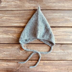 以下が含まれることがあります： 尖ったトップと長いネクタイが付いた灰色のニット帽。帽子は木製の表面の上に置かれています