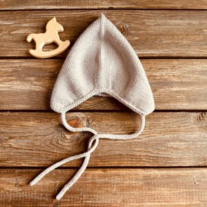 May include: A light gray knitted baby bonnet with tie strings, resting on a wooden surface. A wooden rocking horse toy is next to the bonnet. The bonnet is made of soft, textured yarn.