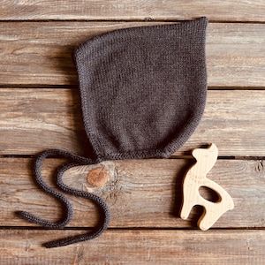 May include: A dark brown knitted bonnet with a rounded shape and two long, thin knitted ties extending from the bottom. Beside the bonnet is a light-colored wooden toy shaped like a stylized animal with a cutout center and visible grain.