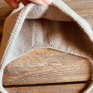 May include: A light beige knitted baby hat with ear flaps and ties. The hat is made of a soft, textured yarn and is shown against a wooden background. The hat is designed to keep a baby's head warm.