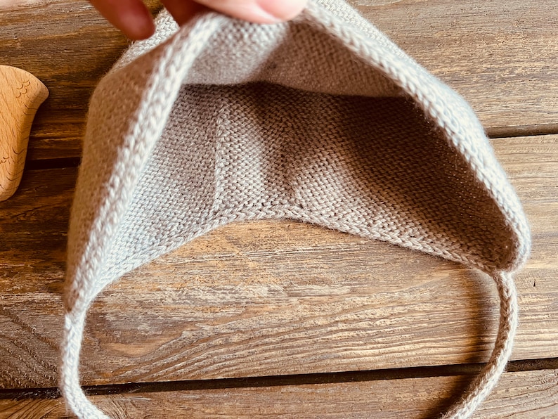 May include: A light beige knitted baby hat with ear flaps and ties. The hat is made of a soft, textured yarn and is shown against a wooden background. The hat is designed to keep a baby's head warm.