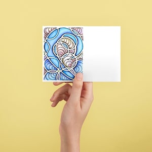 May include: A white greeting card held against a yellow background. The card features a colorful abstract design with blue, pink, and white swirling patterns. The card is held by a hand.