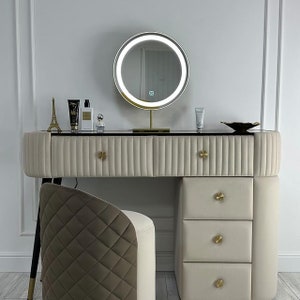 Pu&ograve; includere: Un tavolo da trucco bianco con uno specchio con bordo dorato e tre cassetti. Il tavolo ha un piano nero e pomelli dorati. Uno sgabello piccolo con un tessuto trapuntato marrone e bianco si trova davanti al tavolo.