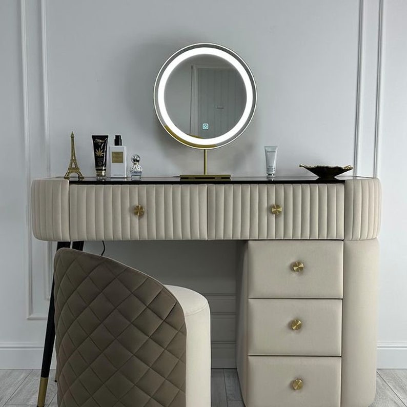 Pu&ograve; includere: Un tavolo da trucco bianco con uno specchio con bordo dorato e tre cassetti. Il tavolo ha un piano nero e pomelli dorati. Uno sgabello piccolo con un tessuto trapuntato marrone e bianco si trova davanti al tavolo.