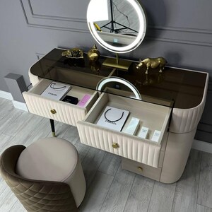 Pu&ograve; includere: Un tavolo da trucco bianco con due cassetti e un piano in vetro. Il tavolo ha una bordatura dorata e una manopola dorata su ogni cassetto. Il tavolo ha uno specchio rotondo dorato con un anello luminoso intorno. Una piccola statuetta di rinoceronte dorata &egrave; sul tavolo. Uno sgabello bianco con una seduta imbottita marrone &egrave; in primo piano.