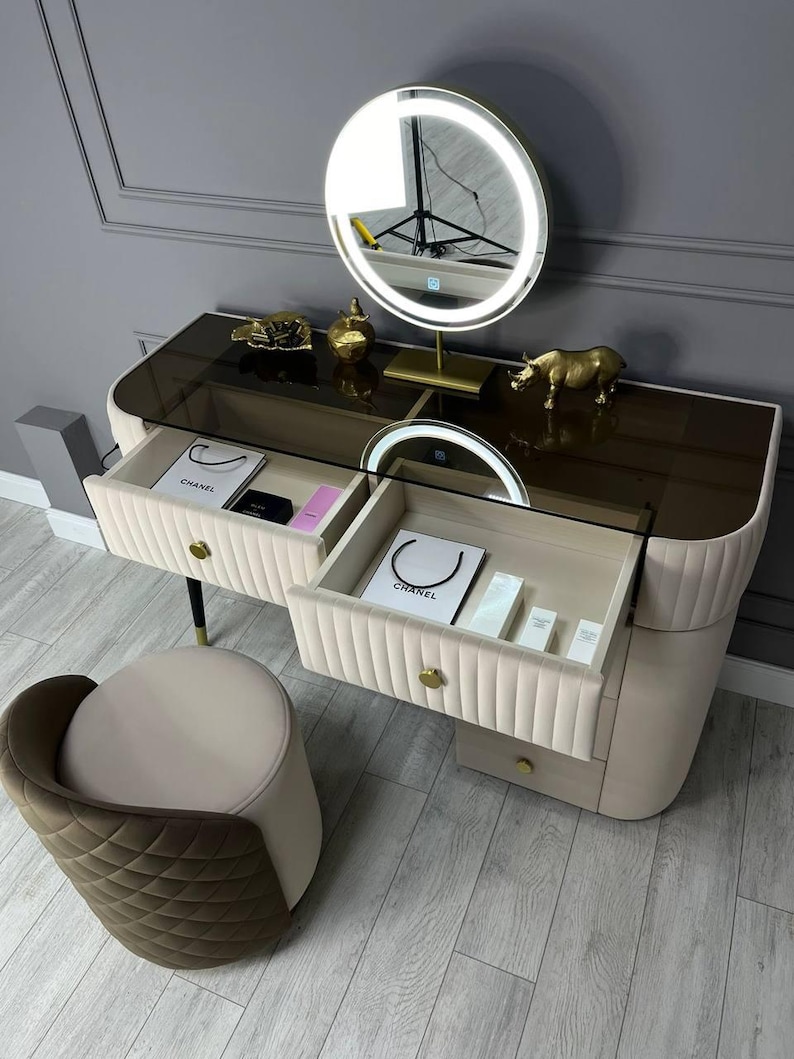 Pu&ograve; includere: Un tavolo da trucco bianco con due cassetti e un piano in vetro. Il tavolo ha una bordatura dorata e una manopola dorata su ogni cassetto. Il tavolo ha uno specchio rotondo dorato con un anello luminoso intorno. Una piccola statuetta di rinoceronte dorata &egrave; sul tavolo. Uno sgabello bianco con una seduta imbottita marrone &egrave; in primo piano.