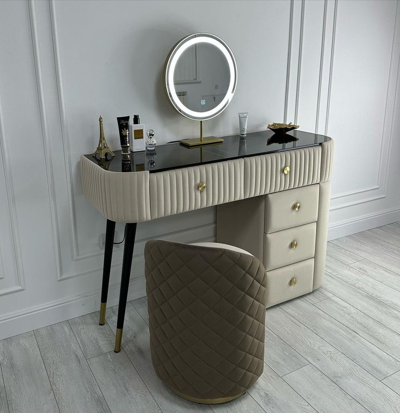 Pu&ograve; includere: Un tavolo da trucco bianco con un piano in vetro nero e dettagli dorati. Il tavolo ha tre cassetti e uno specchio rotondo con luci. Uno sgabello imbottito marrone si trova davanti al tavolo.