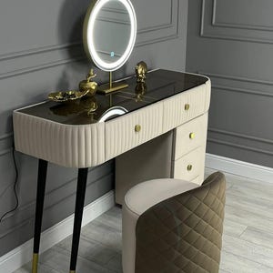 Pu&ograve; includere: Un tavolo da trucco bianco con un piano in vetro e dettagli dorati. Il tavolo ha tre cassetti e quattro gambe nere con punte dorate. Uno specchio rotondo illuminato si trova sul tavolo. Uno sgabello imbottito marrone e bianco si trova di fronte al tavolo.