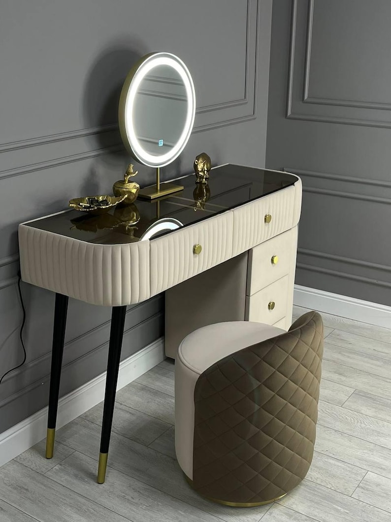 Pu&ograve; includere: Un tavolo da trucco bianco con un piano in vetro e dettagli dorati. Il tavolo ha tre cassetti e quattro gambe nere con punte dorate. Uno specchio rotondo illuminato si trova sul tavolo. Uno sgabello imbottito marrone e bianco si trova di fronte al tavolo.