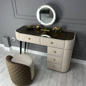 Pu&ograve; includere: Un tavolo da trucco bianco con un piano in vetro marrone e dettagli dorati. Il tavolo ha quattro cassetti e uno specchio rotondo con un anello di luce bianco. Uno sgabello piccolo con un tessuto trapuntato marrone e bianco si trova davanti al tavolo.