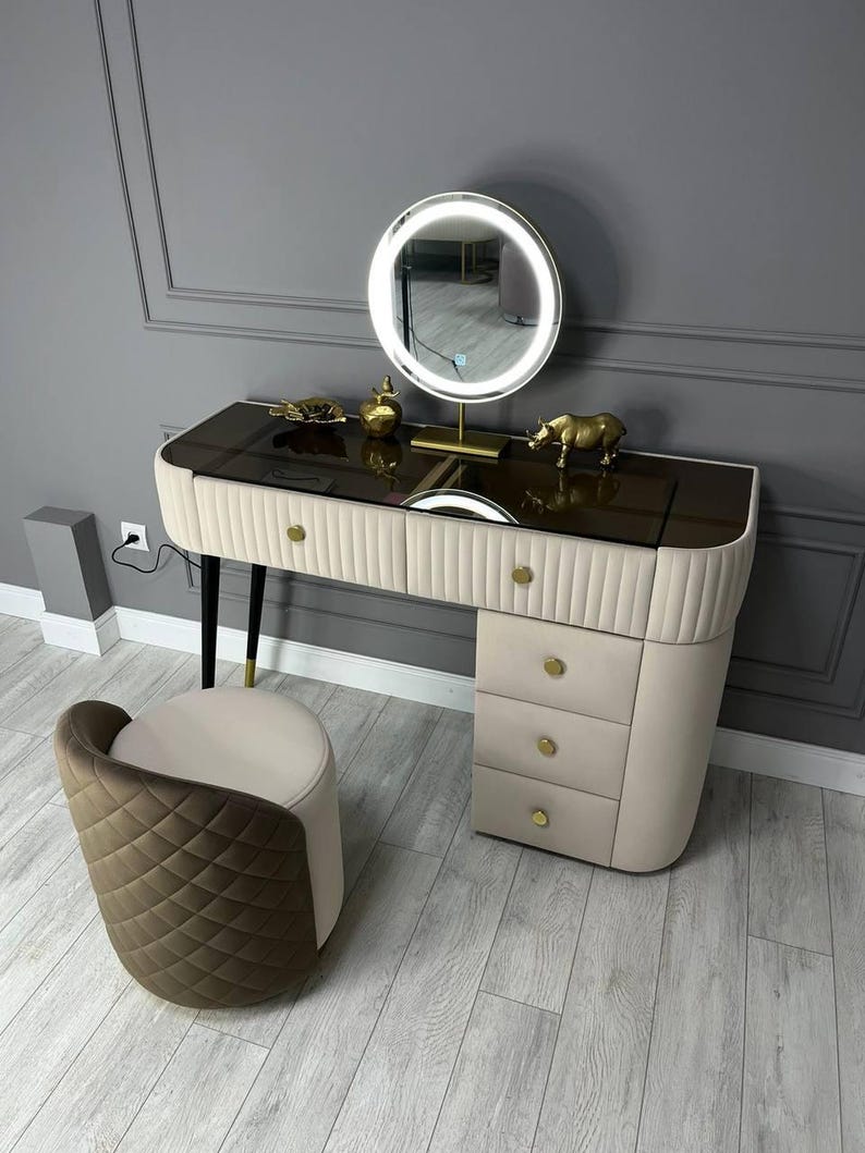 Pu&ograve; includere: Un tavolo da trucco bianco con un piano in vetro marrone e dettagli dorati. Il tavolo ha quattro cassetti e uno specchio rotondo con un anello di luce bianco. Uno sgabello piccolo con un tessuto trapuntato marrone e bianco si trova davanti al tavolo.