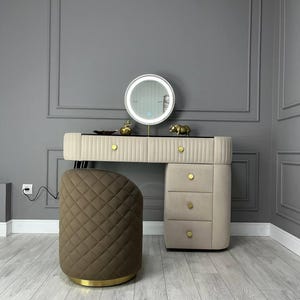 Pu&ograve; includere: Un mobile da bagno bianco con uno specchio rotondo e tre cassetti. Il mobile da bagno ha dettagli dorati e uno sgabello imbottito marrone.
