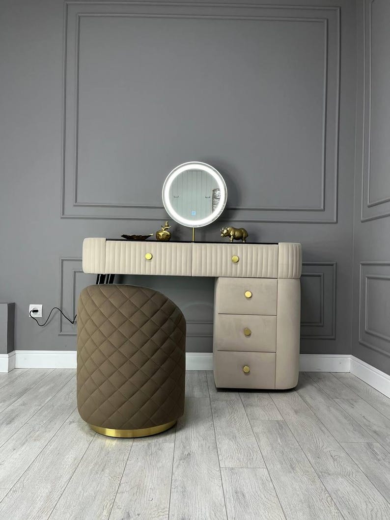 Pu&ograve; includere: Un mobile da bagno bianco con uno specchio rotondo e tre cassetti. Il mobile da bagno ha dettagli dorati e uno sgabello imbottito marrone.