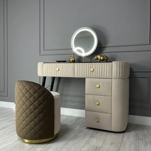 Pu&ograve; includere: Un set da trucco con un mobile da trucco imbottito beige con tre cassetti e uno sgabello imbottito marrone con finiture dorate. Il mobile da trucco ha uno specchio rotondo con un anello luminoso intorno. Il mobile da trucco &egrave; davanti a un muro grigio.