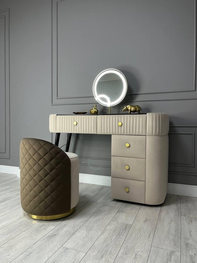 Pu&ograve; includere: Un set da trucco con un mobile da trucco imbottito beige con tre cassetti e uno sgabello imbottito marrone con finiture dorate. Il mobile da trucco ha uno specchio rotondo con un anello luminoso intorno. Il mobile da trucco &egrave; davanti a un muro grigio.