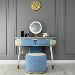 May include: A white vanity table with a blue velvet top and gold trim. The table has a drawer and two legs. A round blue velvet stool with gold trim sits in front of the table. A round mirror with a white halo light is mounted on the wall above the table.