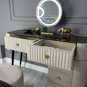 Pu&ograve; includere: Un tavolo da trucco bianco con un piano in vetro e dettagli dorati. Il tavolo ha quattro cassetti e uno specchio rotondo con cornice dorata e luci LED. Una statuetta di rinoceronte dorata &egrave; sul tavolo.
