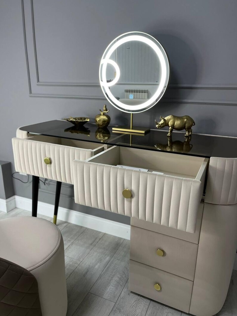 Pu&ograve; includere: Un tavolo da trucco bianco con un piano in vetro e dettagli dorati. Il tavolo ha quattro cassetti e uno specchio rotondo con cornice dorata e luci LED. Una statuetta di rinoceronte dorata &egrave; sul tavolo.