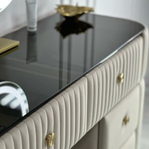 Pu&ograve; includere: Un tavolo da trucco con un piano in vetro nero e cassetti imbottiti beige con pomelli dorati. I cassetti hanno un design a coste verticali.