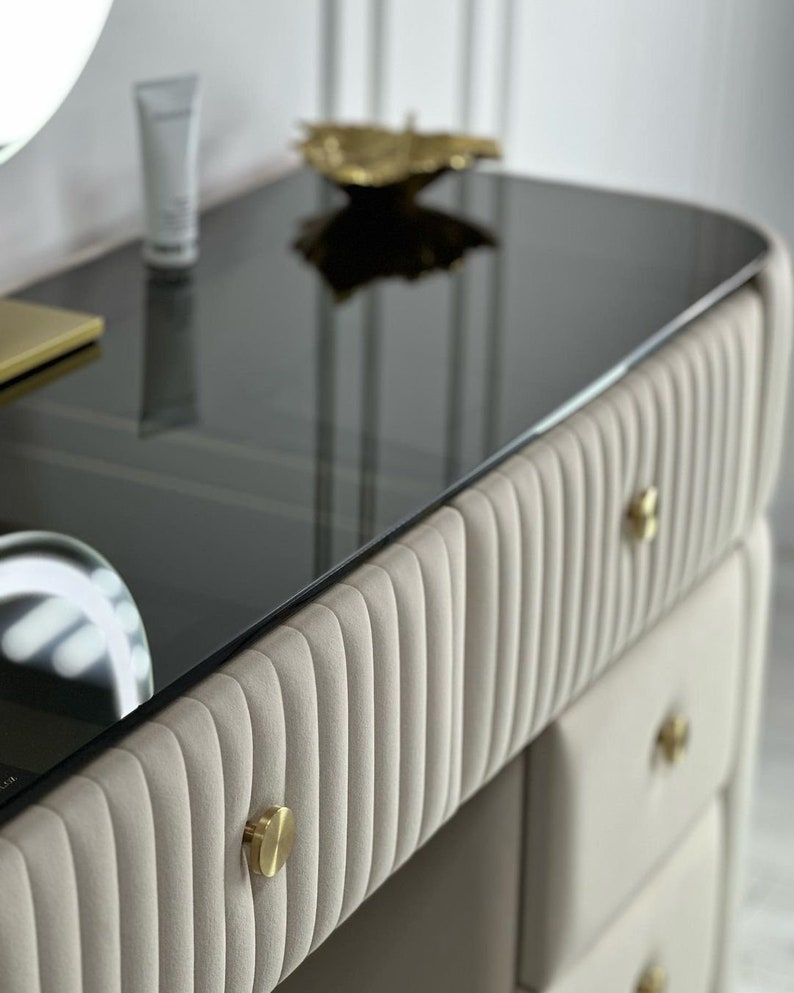 Pu&ograve; includere: Un tavolo da trucco con un piano in vetro nero e cassetti imbottiti beige con pomelli dorati. I cassetti hanno un design a coste verticali.