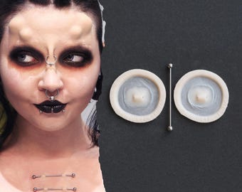 Könnte beinhalten: Zwei Brustwarzenabdeckungen mit einem Barbell-Piercing auf dunklem Hintergrund. Die Brustwarzenabdeckungen sind weiß und kreisförmig. Die Langhantel ist silberfarben. Das Bild zeigt auch eine Person mit Gesichtspiercings und Körpermodifikationen.