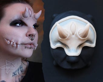 Könnte beinhalten: Eine Person mit künstlichen Hörnern und Zähnen sowie einer passenden Maske. Die Hörner und Zähne sind hellrosa, und die Maske ist schwarz mit einem weißen und hellrosa Horndesign. Die Person hat mehrere Gesichtspiercings und dunkles Augen-Make-up.