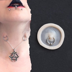 May include: A silver-colored body jewelry set featuring a pendant necklace with a biohazard symbol, connected to two circular piercings. A separate image shows a silver captive bead ring. The jewelry is displayed on a black background.