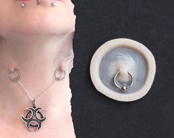 Könnte beinhalten: Silberfarbenes Körperschmuck-Set. Das Set beinhaltet eine Halskette mit Biohazard-Symbol, verbunden mit zwei kreisförmigen Piercings. Ein separates Bild zeigt einen silbernen Klemmkugelring. Schwarzer Hintergrund.