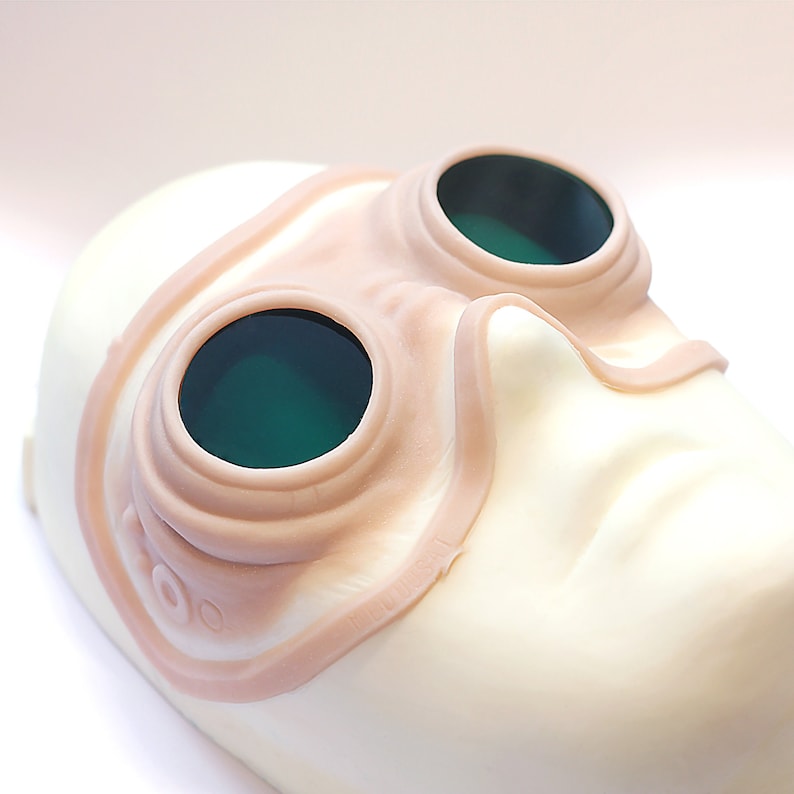 Steampunk silicone prosthetics googles in vanilla shade with green lenses on a white surface