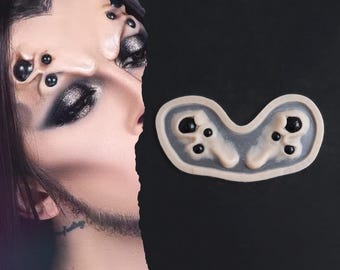 Puede incluir: Una prótesis de silicona con una base de tono piel pálido, múltiples protuberancias carnosas en relieve y esferas negras brillantes incrustadas. La pieza está diseñada para aplicarse en la frente y crear un efecto de criatura con varios ojos.