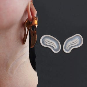 May include: Gold-colored, abstract earrings with a unique, curved design are shown in this image. One earring is worn on an ear, while two matching earrings are displayed separately. The earrings have a smooth, polished finish and a modern aesthetic.