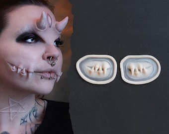 Könnte beinhalten: Eine Person mit künstlichen Hörnern und Fangzähnen, die ein theatralisches Make-up zeigt. Zwei Sätze künstlicher Fangzähne werden in ihren Formen gezeigt. Das Make-up umfasst dunklen Lidschatten und Gesichtspiercings, was einen dramatischen Effekt erzeugt.