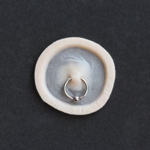 May include: A silver ring with a small ball on the end, sitting on a white circular surface with the words "Genius Studio" printed on it.