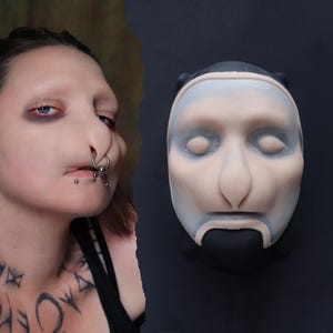 May include: A pale-coloured mask with a human face, featuring a prominent nose and closed eyes. The mask has a black base and a light blue wash on the forehead and cheeks. The mask is displayed next to a person with facial piercings.