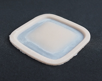 May include: A square-shaped silicone mold with rounded corners, featuring a raised beige border and a recessed, smooth, light-colored center. The border has embossed text that reads "THE DUSAT STUDIO" along one of its edges.