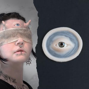 May include: A surreal image featuring a person with multiple piercings, a blindfold, and an eye on their forehead. A circular, eye-shaped art piece is on the right. The art piece has a light blue and white border with a detailed eye in the center.